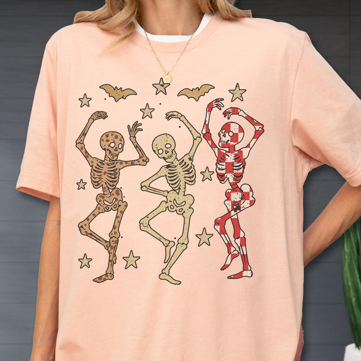May include: A light peach-colored t-shirt featuring a graphic of three dancing skeletons. The skeletons are surrounded by stars and bats. One skeleton is red and white checkered. The shirt has a white collar.