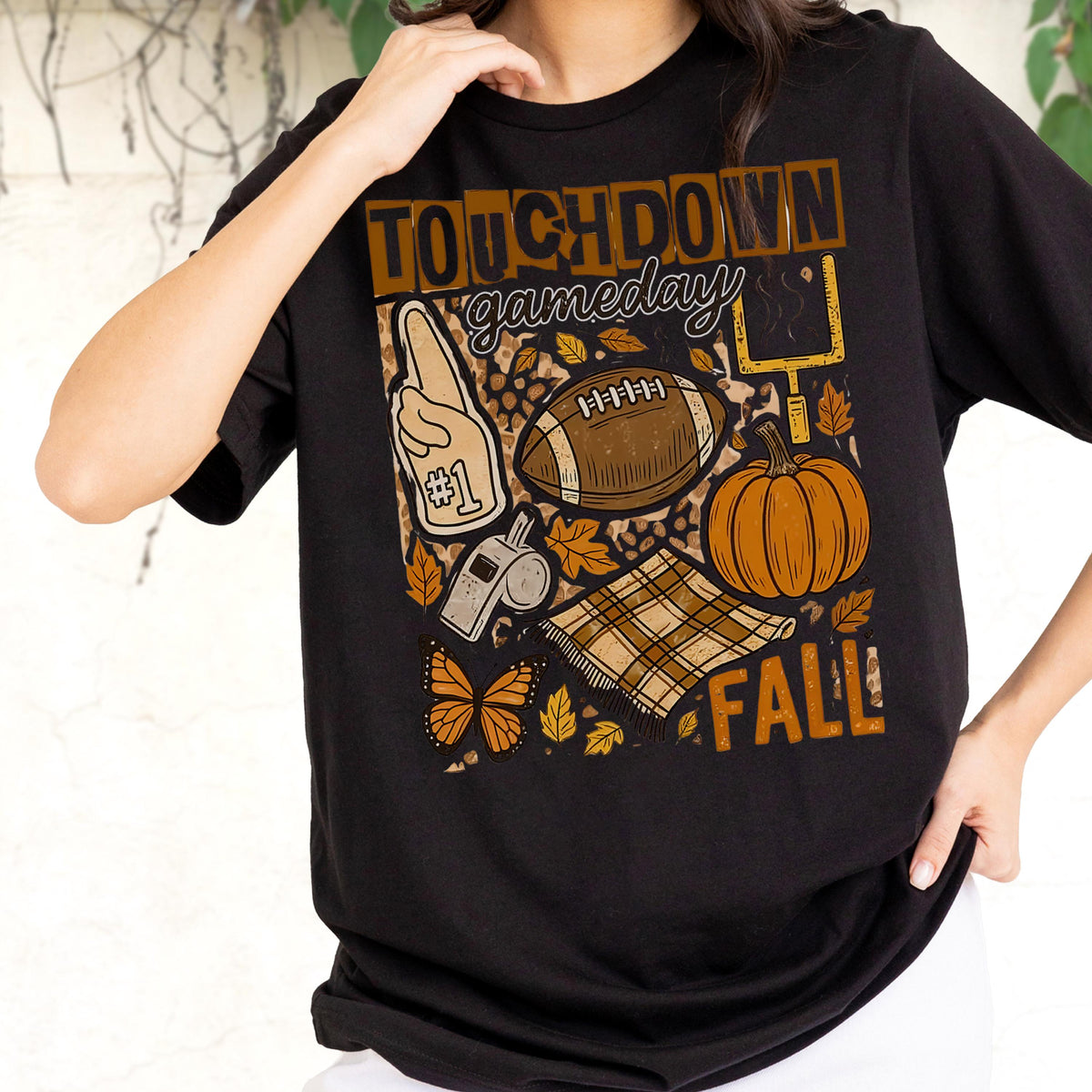 May include: Black t-shirt with a fall-themed graphic. The design includes a football, pumpkin, and the words "TOUCHDOWN gameday FALL". Other elements include a foam finger, whistle, and plaid blanket.