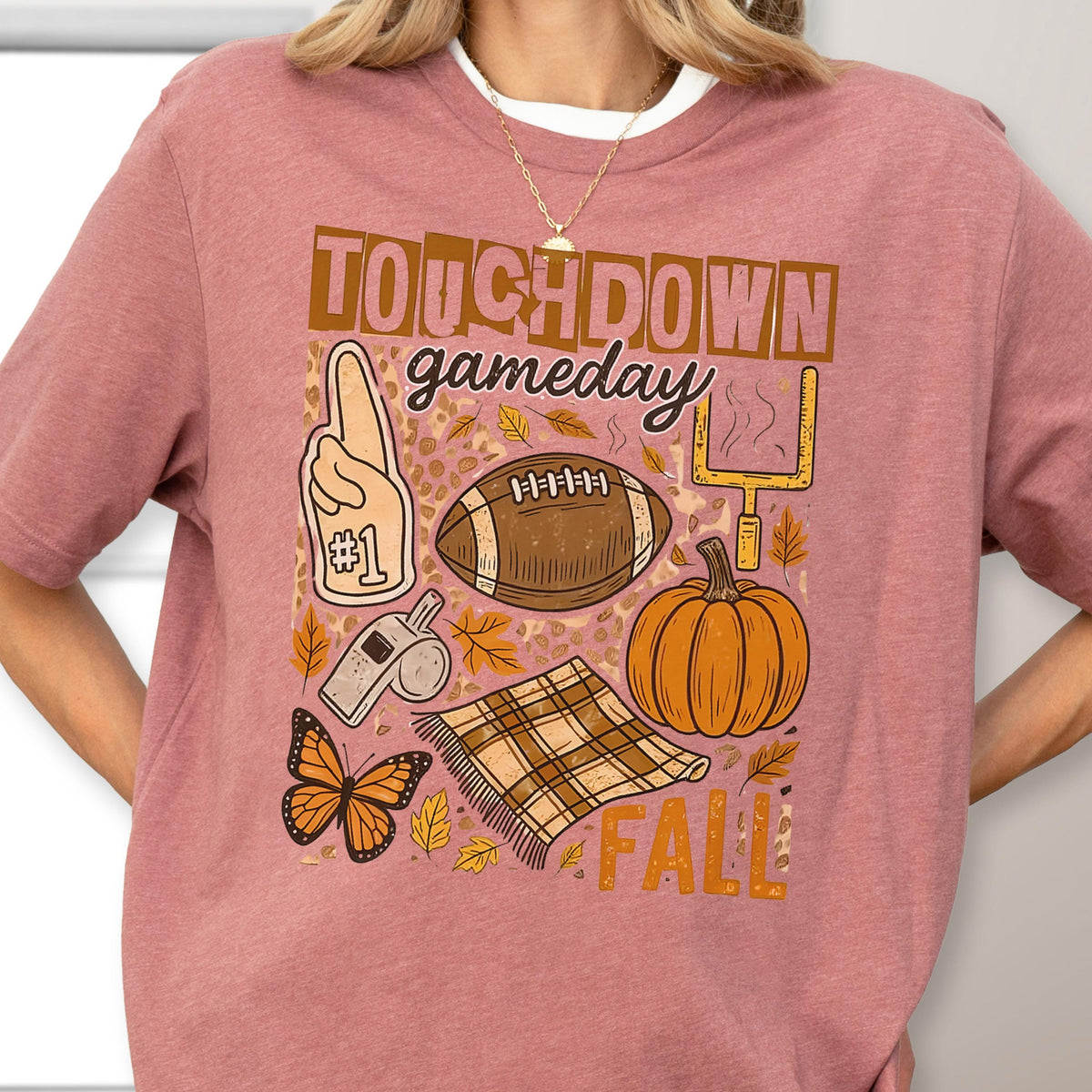May include: A mauve-colored t-shirt with a graphic design featuring the words "Touchdown Gameday Fall". The design includes a football, pumpkin, foam finger, whistle, butterfly, and a plaid blanket.