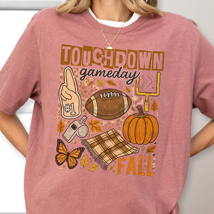 May include: A mauve-colored t-shirt with a graphic design featuring the words "Touchdown Gameday Fall". The design includes a football, pumpkin, foam finger, whistle, butterfly, and a plaid blanket.