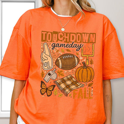 May include: Orange t-shirt with a "TOUCHDOWN gameday FALL" graphic. The design includes a foam finger, football, pumpkin, goalpost, whistle, butterfly, and a plaid blanket. The shirt is a crew neck.