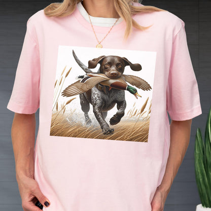 May include: Pink t-shirt featuring a graphic of a hunting dog with a duck in its mouth, running through tall grass. The dog is brown and white, and the duck has brown, green, and white feathers. The shirt has short sleeves and a crew neck.