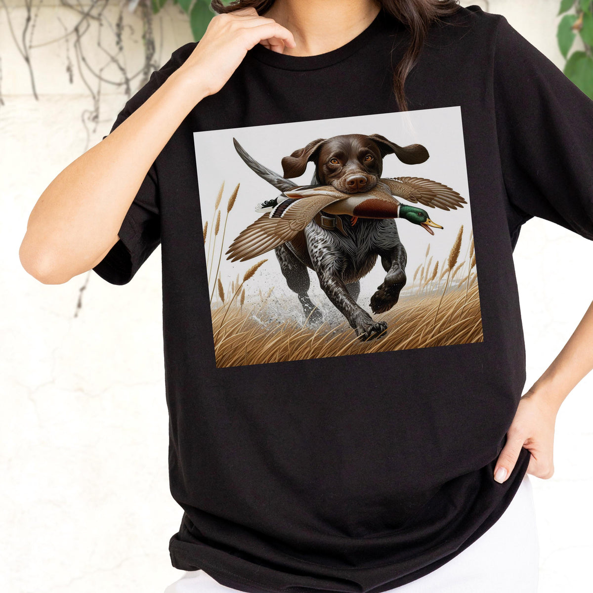 May include: Black t-shirt featuring a square graphic of a hunting dog with a duck in its mouth, running through tall grass. The dog is brown and white with floppy ears. The duck has brown and green feathers. The background is a light gray.