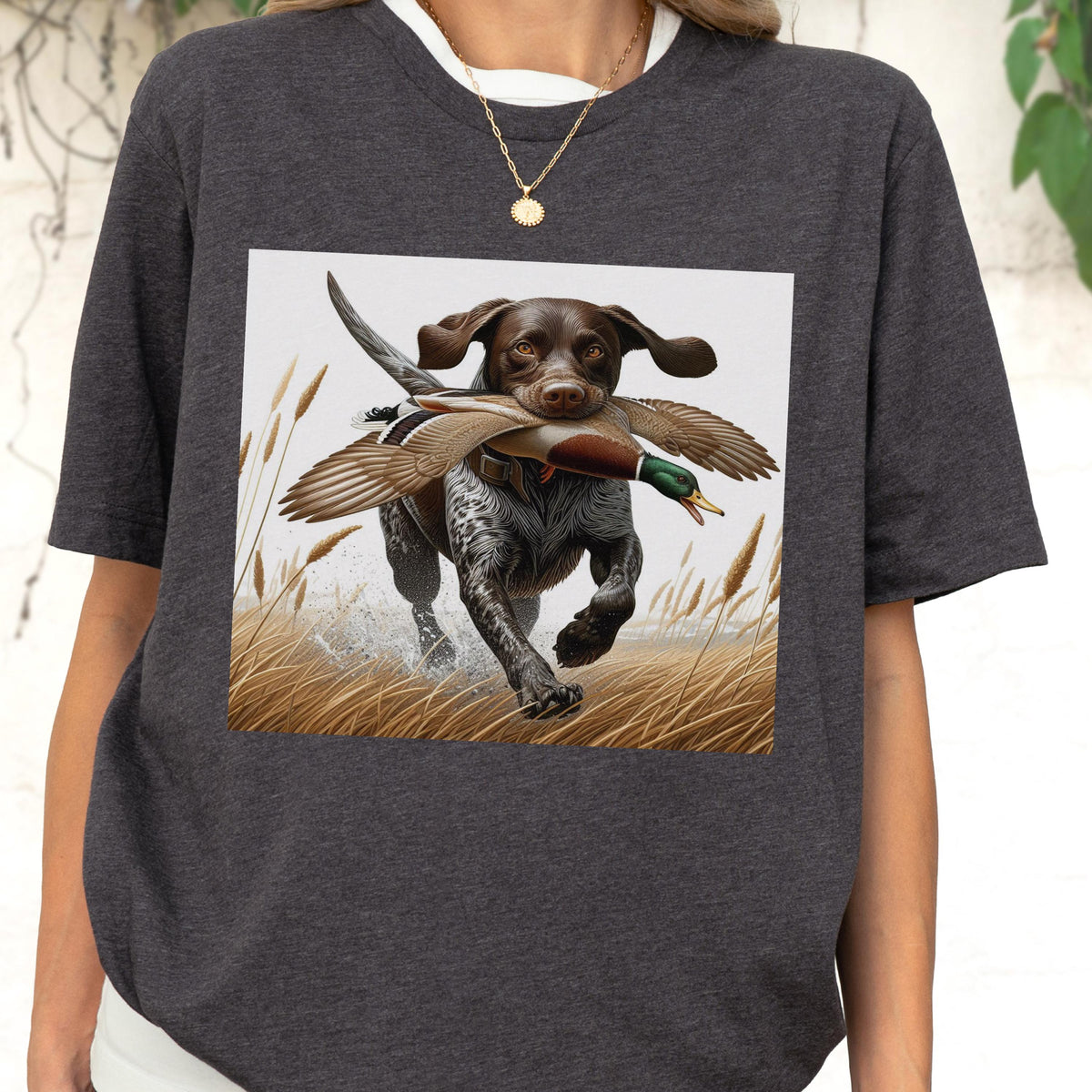 May include: Dark gray t-shirt featuring a graphic of a hunting dog with brown and white fur, carrying a duck in its mouth. The dog is running through tall grass with water splashing around its feet. The image has a vintage, outdoorsy feel.