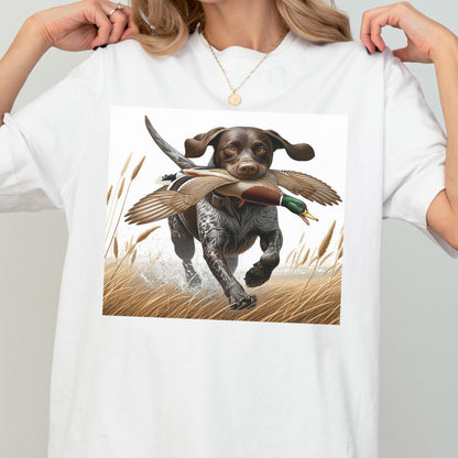 May include: White t-shirt featuring a digital illustration of a hunting dog with brown and white fur, running through tall grass while carrying a duck in its mouth. The dog is in motion, with water splashing around its paws.