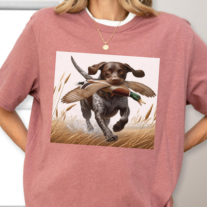 May include: A dusty rose t-shirt featuring a graphic of a brown and white dog running through tall grass, carrying a duck in its mouth. The dog is the focal point, with water splashing around its paws. The shirt has a crew neck.