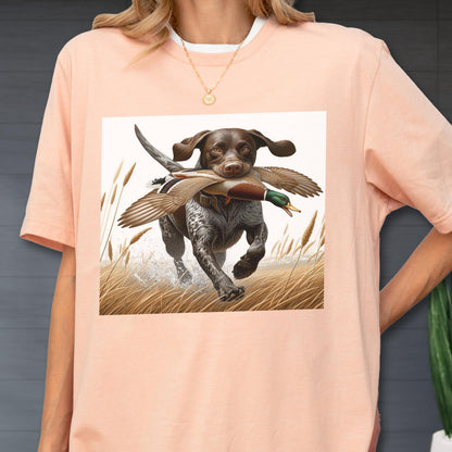 May include: A light peach-colored t-shirt featuring a graphic of a hunting dog with a duck in its mouth, running through tall grass. The dog is brown and white, and the duck has green and brown feathers. The shirt has a white collar.