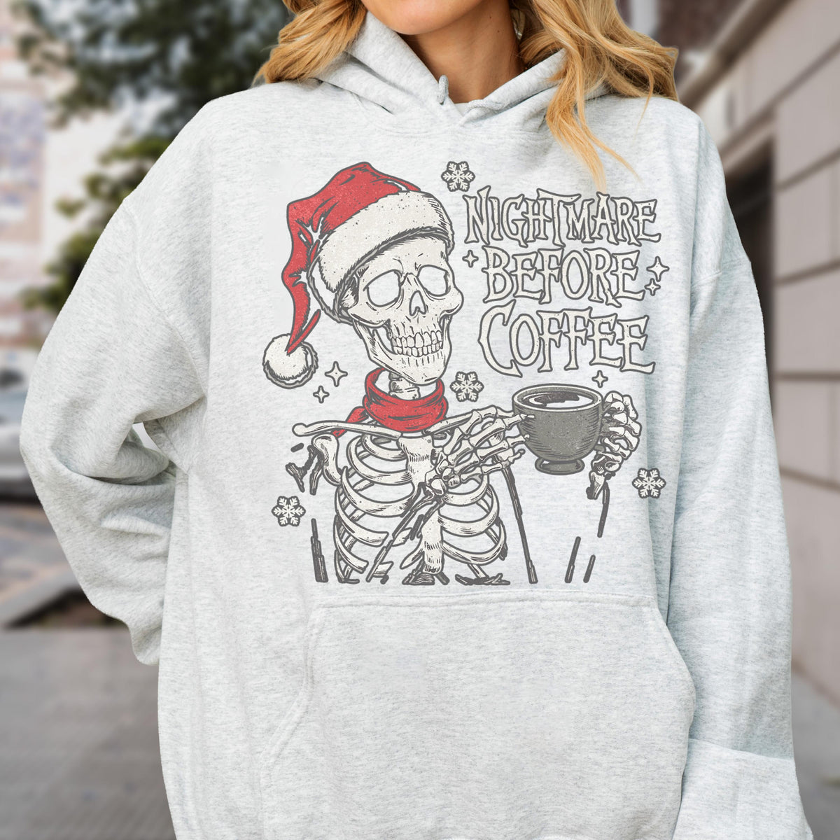 May include: Light gray hoodie featuring a skeleton wearing a Santa hat and red scarf, holding a coffee cup. The text "NIGHTMARE BEFORE COFFEE" is printed above the skeleton. The design includes snowflakes.