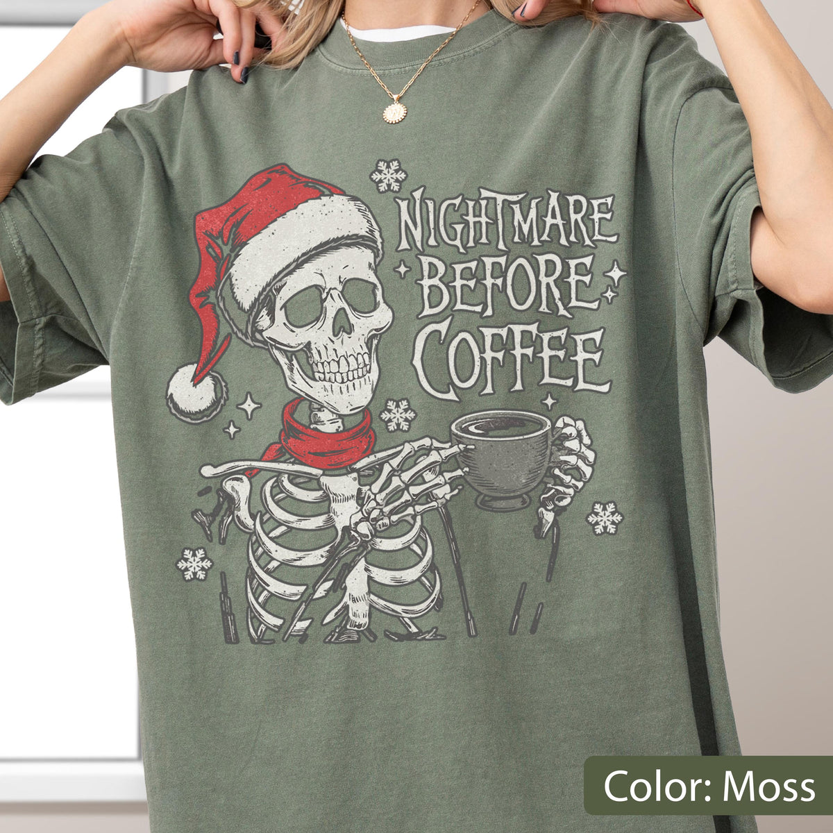 May include: Moss green t-shirt featuring a skeleton wearing a Santa hat and red scarf, holding a coffee cup. The text "NIGHTMARE BEFORE COFFEE" is printed above the skeleton. Snowflake graphics are scattered around the design.