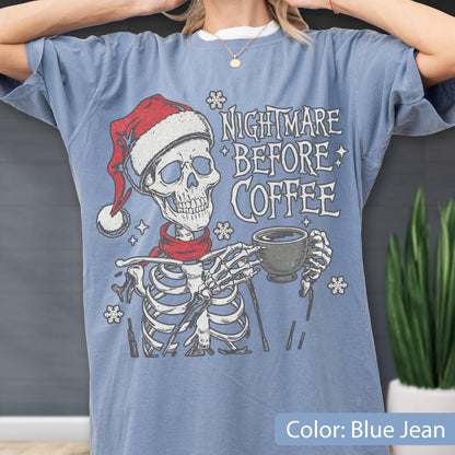 May include: Blue jean-colored t-shirt featuring a skeleton wearing a Santa hat and red scarf, holding a coffee cup. The shirt has the text "Nightmare Before Coffee" in white. The shirt is a relaxed fit.