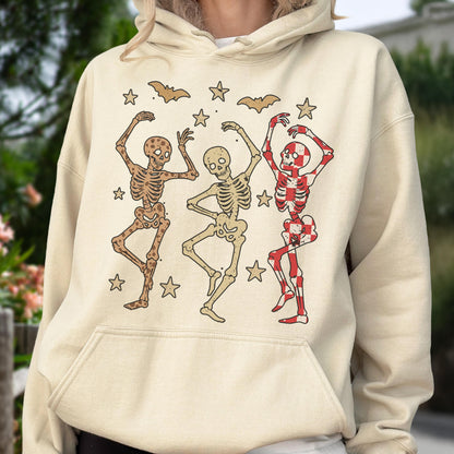 May include: Beige hoodie featuring a graphic of three dancing skeletons in brown and red, with stars and bats. The skeletons are in various poses, with one in a red and white checkered pattern. A fun Halloween-themed design.
