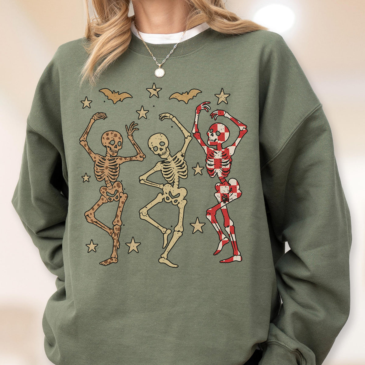 May include: Sage green sweatshirt featuring a graphic of three dancing skeletons. The skeletons are in various poses, with one in red and white checkered patterns. Bats and stars are also included in the design.