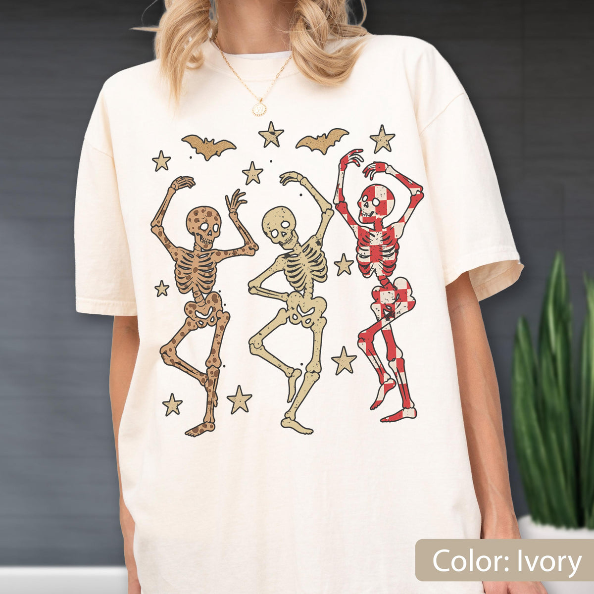 May include: Ivory-colored t-shirt featuring three dancing skeletons in various colors, surrounded by stars and bats. The skeletons have different patterns and colors, with one in red and white checkered design. The shirt is a relaxed fit.
