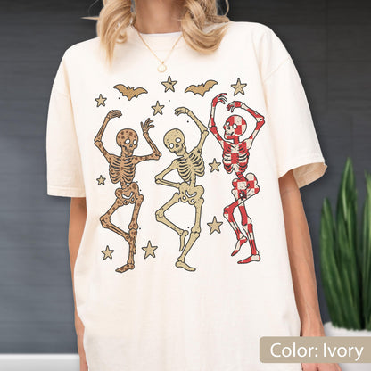 May include: Ivory-colored t-shirt featuring three dancing skeletons in various colors, surrounded by stars and bats. The skeletons have different patterns and colors, with one in red and white checkered design. The shirt is a relaxed fit.