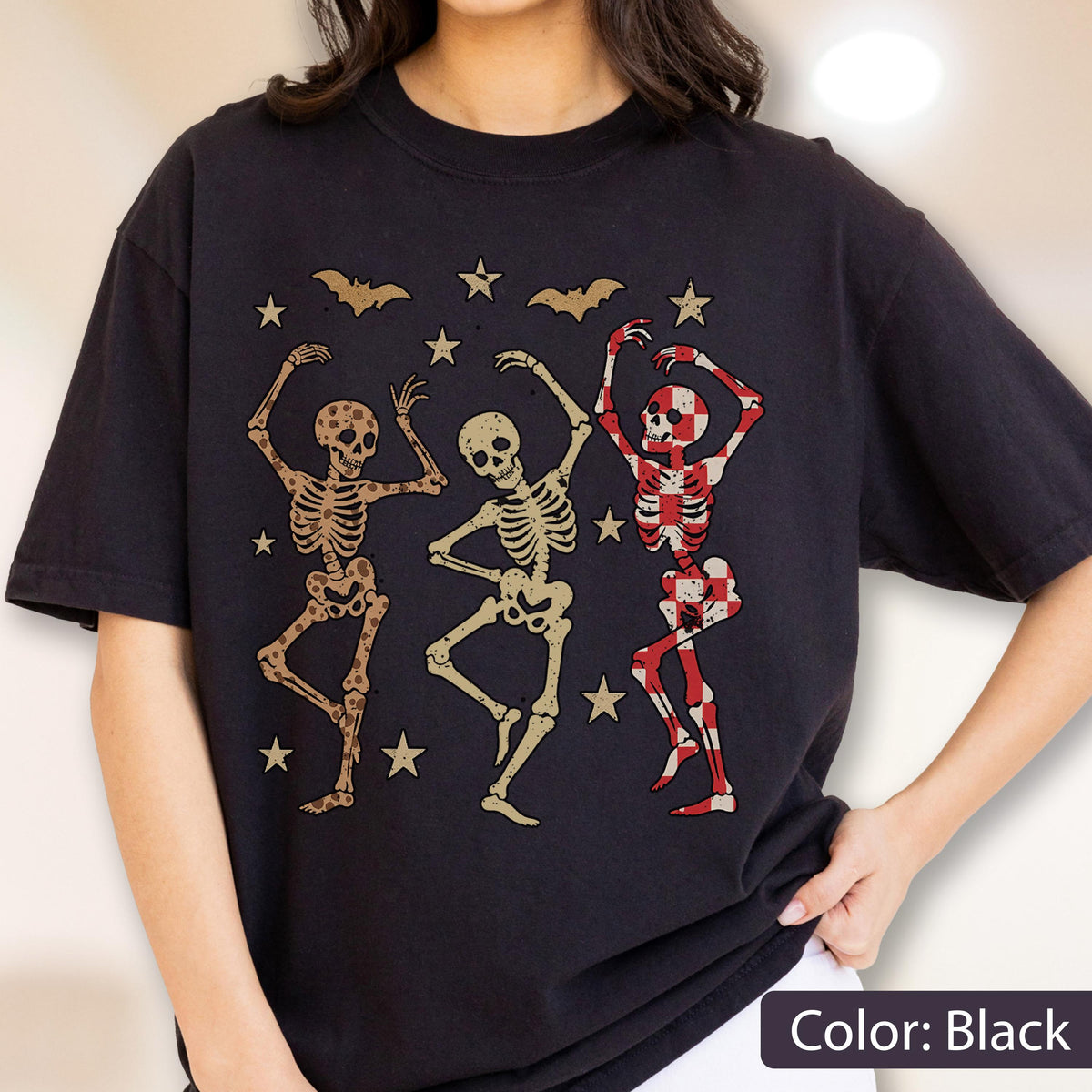 May include: Black t-shirt featuring a graphic of three dancing skeletons, bats, and stars. One skeleton has a red and white checkered pattern. The design is in shades of brown, beige, and red.