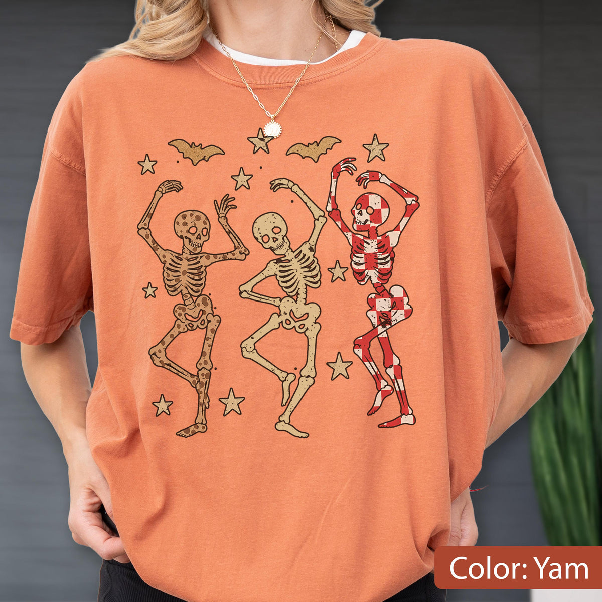 May include: Orange-colored t-shirt featuring three dancing skeleton figures in various poses. The skeletons are surrounded by star and bat graphics. The shirt's color is described as "Yam".