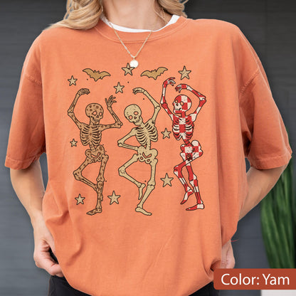 May include: Orange-colored t-shirt featuring three dancing skeleton figures in various poses. The skeletons are surrounded by star and bat graphics. The shirt's color is described as "Yam".