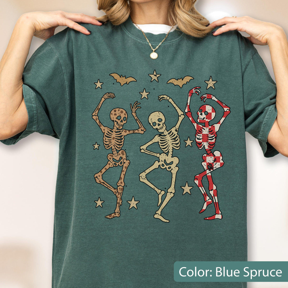 May include: Blue spruce t-shirt featuring a graphic of three dancing skeletons, stars, and bats. The skeletons are in various colors, including brown and red checkered patterns. The shirt is a comfortable, casual style.