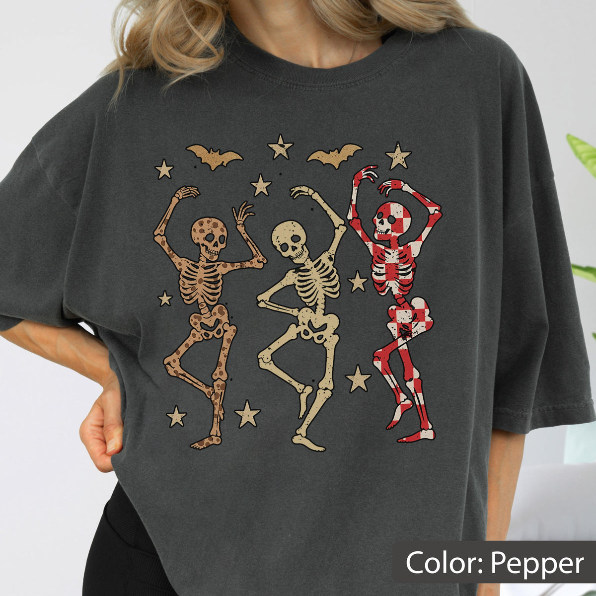 May include: Dark gray t-shirt featuring a graphic of three dancing skeletons in various colors. Bats and stars are scattered around the skeletons. The text "Color: Pepper" is at the bottom right.