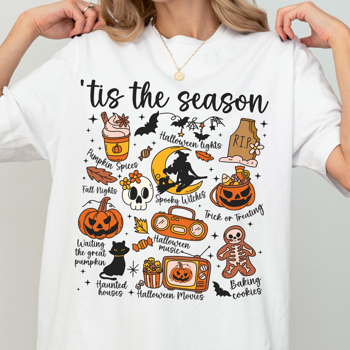 May include: White t-shirt with a colorful Halloween-themed graphic. The design includes pumpkins, bats, a skull, and the phrase "'tis the season." Other text includes "Pumpkin Spices," "Spooky Witches," and "Trick or Treating."