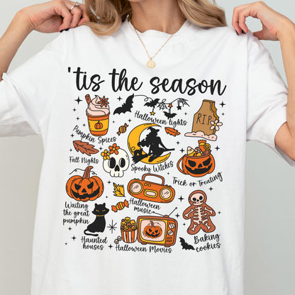 May include: White t-shirt with a colorful Halloween-themed graphic. The design includes pumpkins, bats, a skull, and the phrase "'tis the season." Other text includes "Pumpkin Spices," "Spooky Witches," and "Trick or Treating."