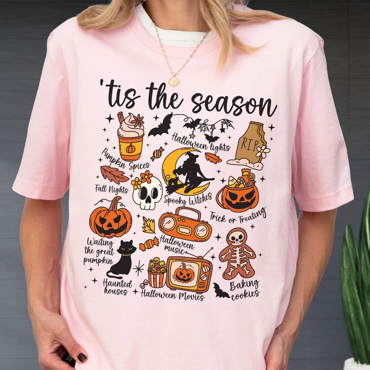 May include: Pink t-shirt with a Halloween-themed graphic. The design includes a pumpkin spice latte, bats, a skull, a black cat, and the phrase "'tis the season". Other elements include a jack-o'-lantern, a witch, and a gingerbread man.
