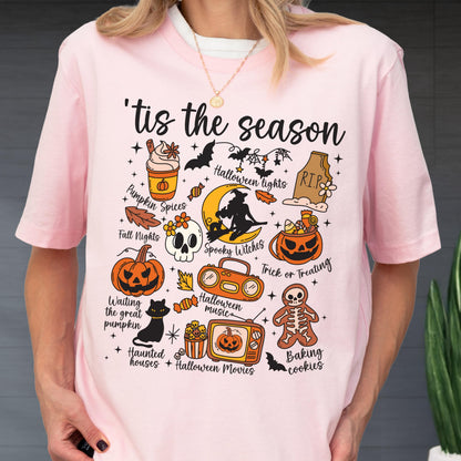 May include: Pink t-shirt with a Halloween-themed graphic. The design includes a pumpkin spice latte, bats, a skull, a black cat, and the phrase "'tis the season". Other elements include a jack-o'-lantern, a witch, and a gingerbread man.