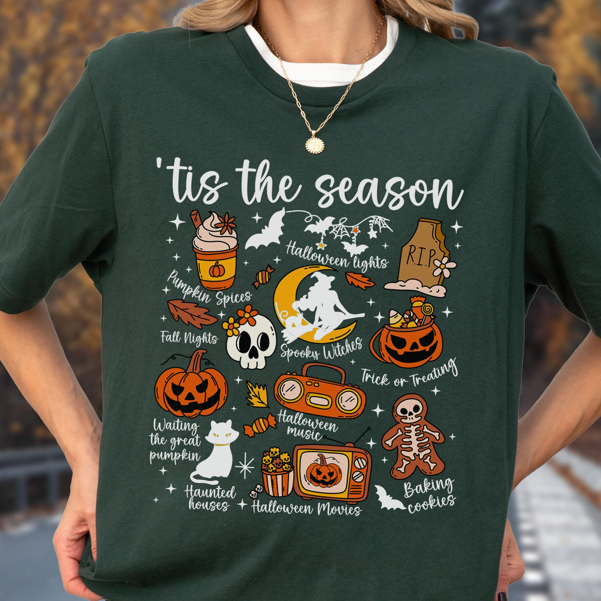 May include: Dark green t-shirt with a festive Halloween design. The shirt features various Halloween-themed illustrations, including pumpkins, bats, and the phrase "'tis the season." Other graphics include a skull, a witch, and the words "Pumpkin Spices" and "Trick or Treating."