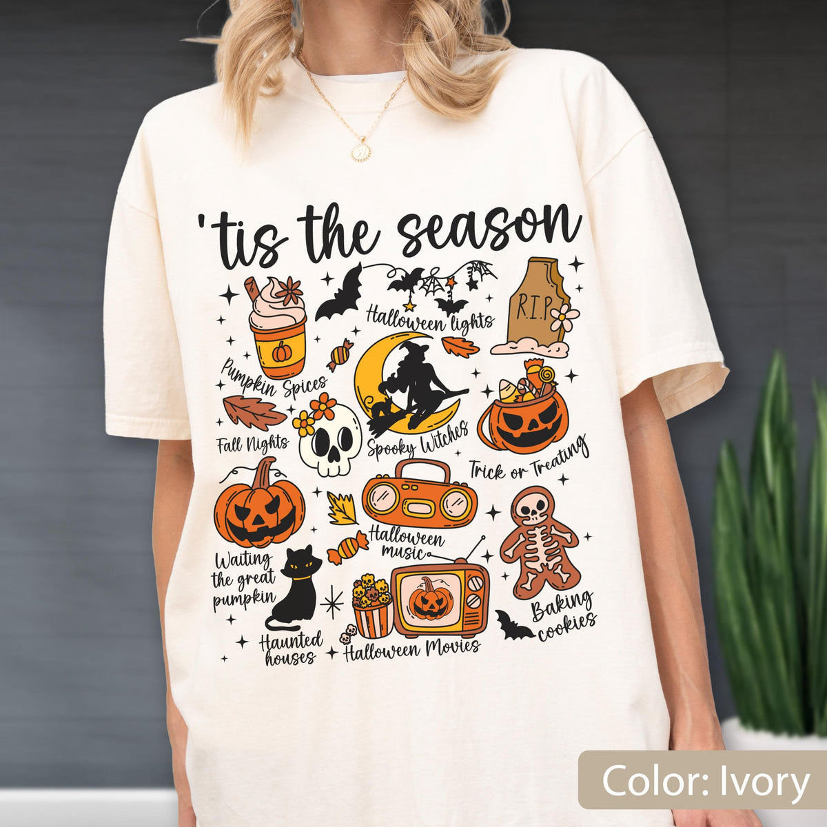 May include: An ivory t-shirt featuring the text 'tis the season and various Halloween-themed illustrations including a pumpkin spice drink, a skull, a jack-o'-lantern, a black cat, a boombox, a television, a gingerbread skeleton, and a witch on a moon.