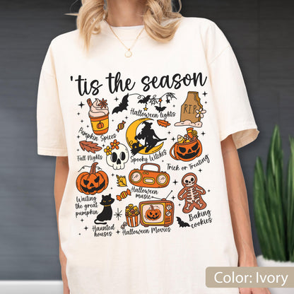 May include: An ivory t-shirt featuring the text 'tis the season and various Halloween-themed illustrations including a pumpkin spice drink, a skull, a jack-o'-lantern, a black cat, a boombox, a television, a gingerbread skeleton, and a witch on a moon.