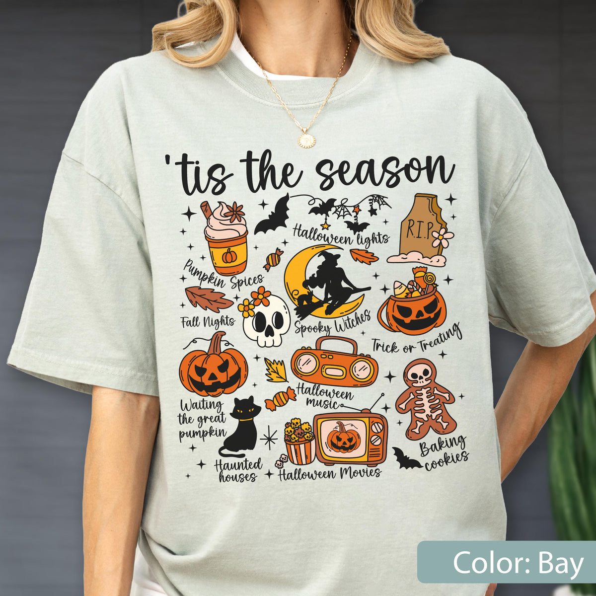 May include: A light bay-colored t-shirt featuring the text 'tis the season and various Halloween-themed illustrations including a pumpkin spice drink, a tombstone, a witch on a moon, a jack-o'-lantern, a skull, a boombox, a television, and a skeleton cookie.
