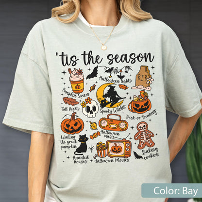 May include: A light bay-colored t-shirt featuring the text 'tis the season and various Halloween-themed illustrations including a pumpkin spice drink, a tombstone, a witch on a moon, a jack-o'-lantern, a skull, a boombox, a television, and a skeleton cookie.