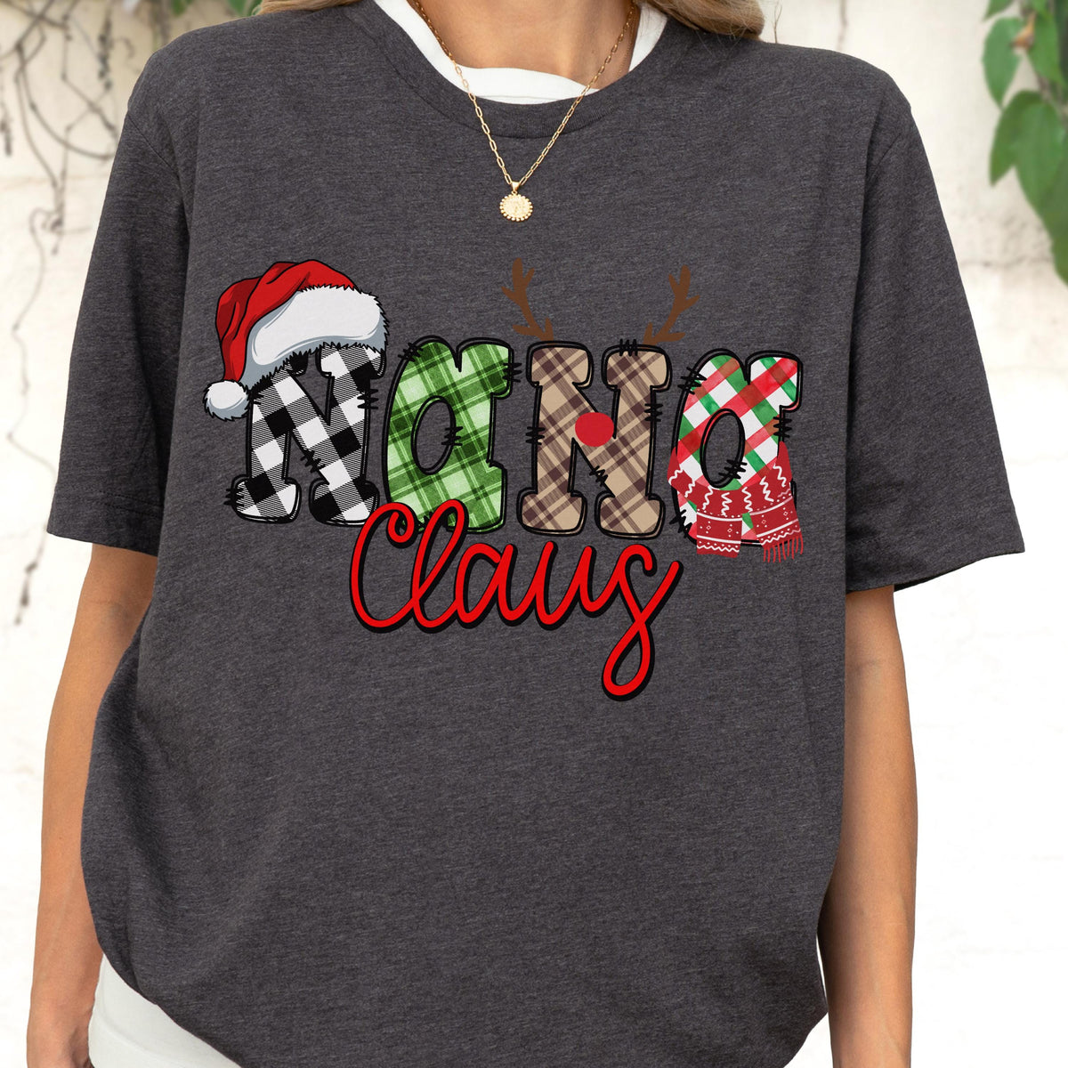 May include: Dark gray t-shirt featuring the text "Nana Claus" in a festive design. The word "Nana" is in a black and white plaid pattern with a Santa hat. The word "Claus" is in red script.