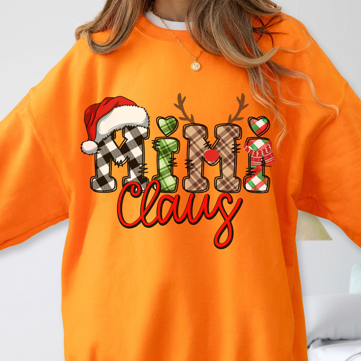 May include: Orange sweatshirt with the text "Nimi Claus" in a festive design. The letters are decorated with a Santa hat, reindeer antlers, hearts, and plaid patterns, perfect for the holiday season.