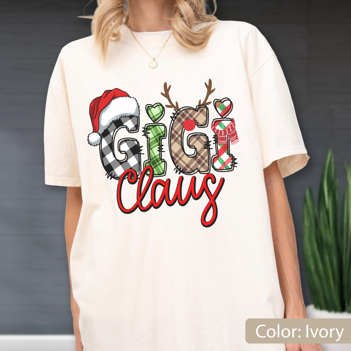 May include: An ivory short-sleeve t-shirt featuring the text GIGI CLAUS in a festive graphic. The letters are filled with black and white buffalo plaid, green plaid, and brown plaid patterns, accented with a red Santa hat, reindeer antlers, and a scarf.