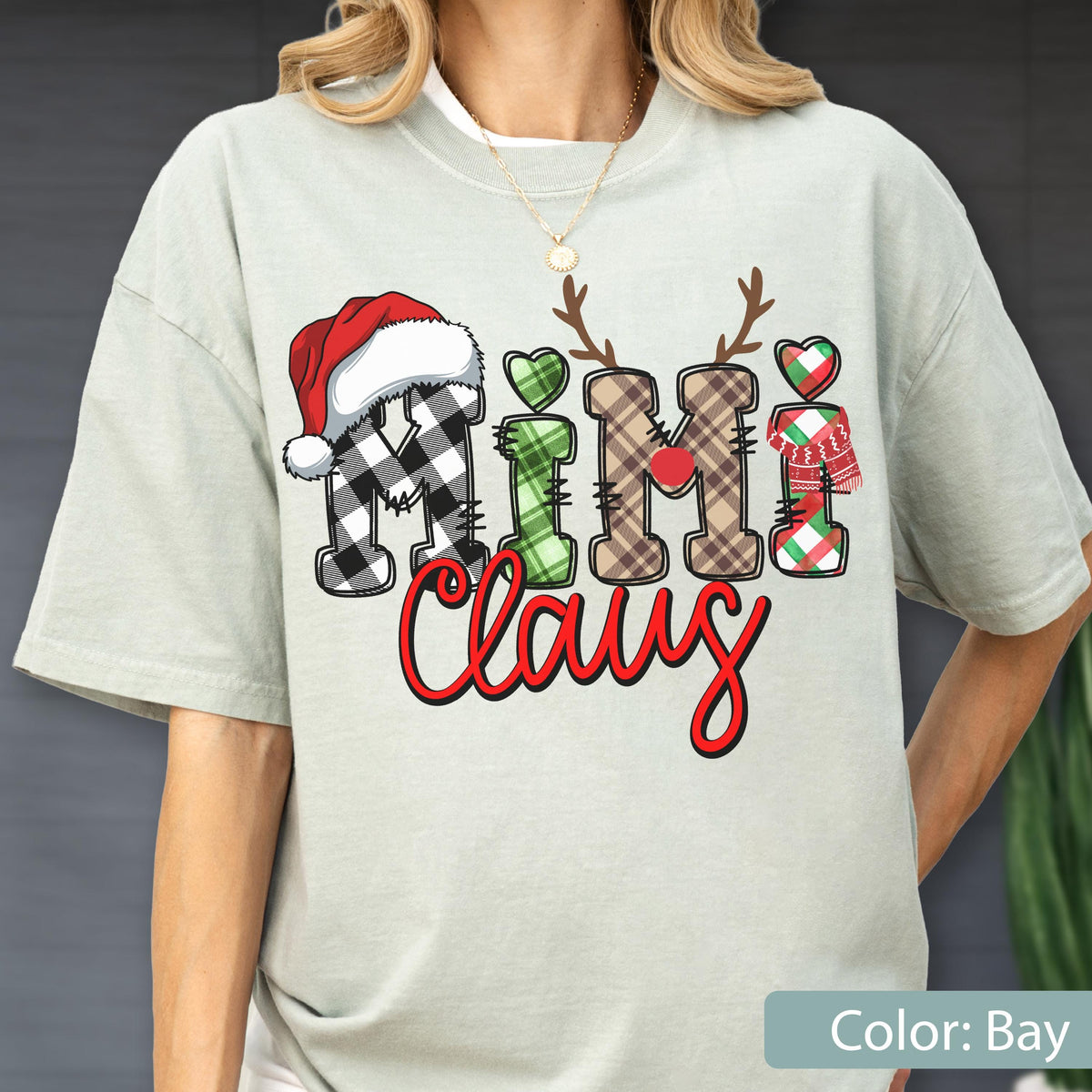 May include: A light sage green t-shirt featuring the text "Mimi Claus" in a decorative font. The letters are styled with plaid patterns, a red Santa hat, reindeer antlers, a red nose, a green heart, and a red and green scarf graphic.