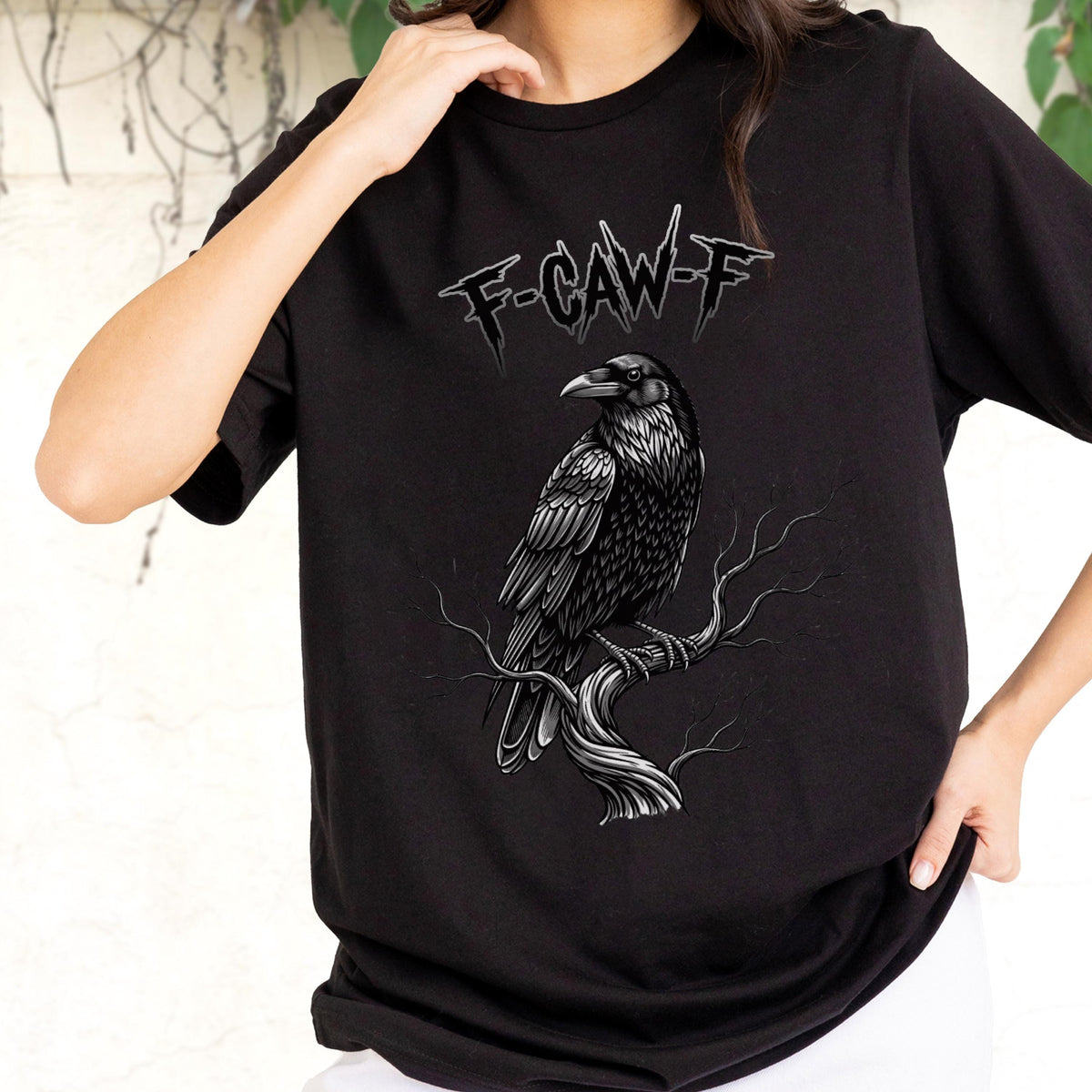 May include: Black t-shirt featuring a detailed grayscale illustration of a raven perched on a branch. The text "F-CAW-F" is printed above the raven in a gothic font. The design is centered on the shirt.