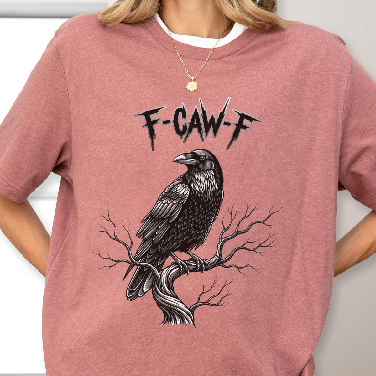 May include: A mauve t-shirt featuring a black raven perched on a tree branch. The raven is detailed with textured feathers. Above the raven is the text "F-CAW-F" in a stylized font. The shirt has short sleeves and a crew neck.