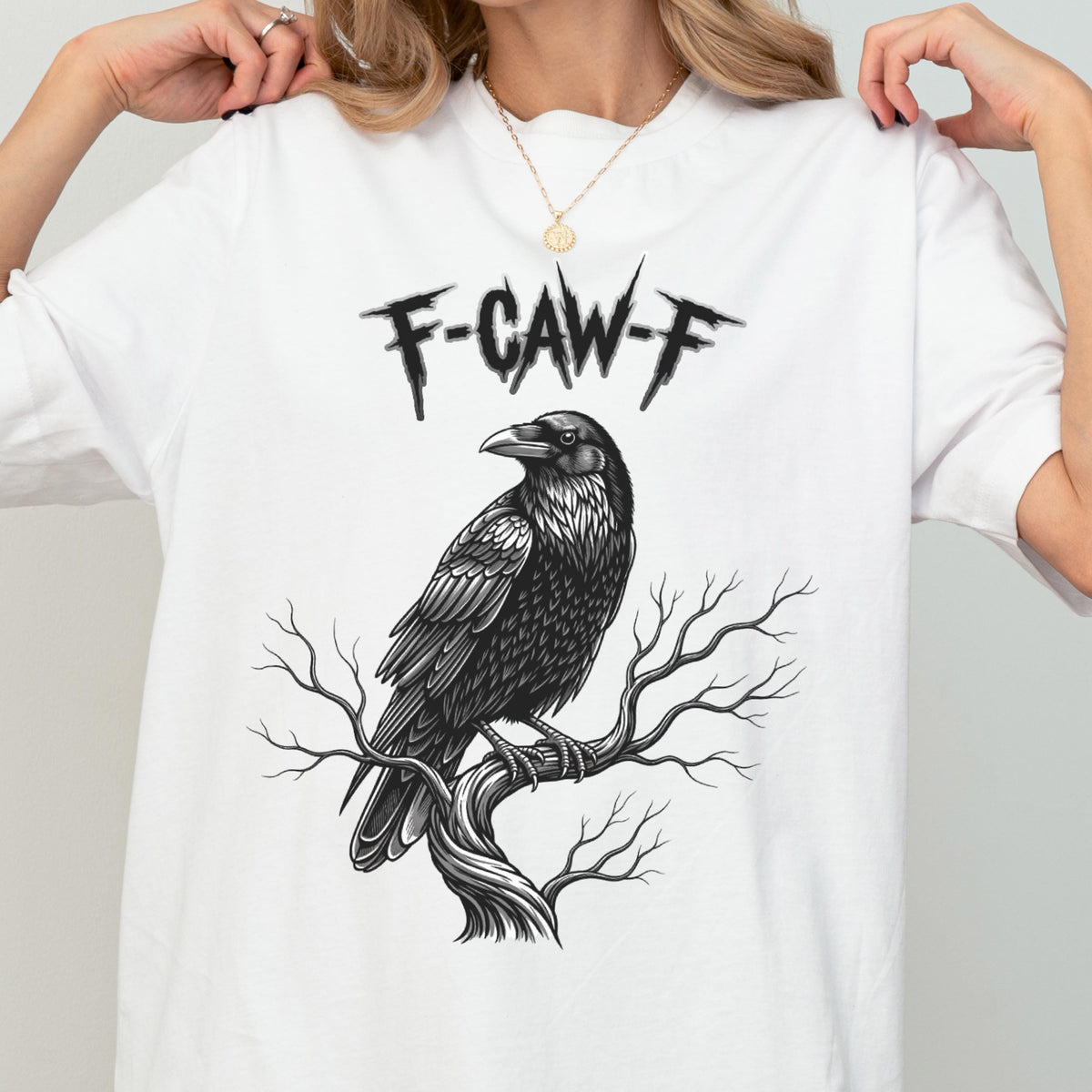 May include: White t-shirt featuring a detailed black raven perched on a tree branch. The text "F-CAW-F" is printed above the raven. The design is a graphic illustration.