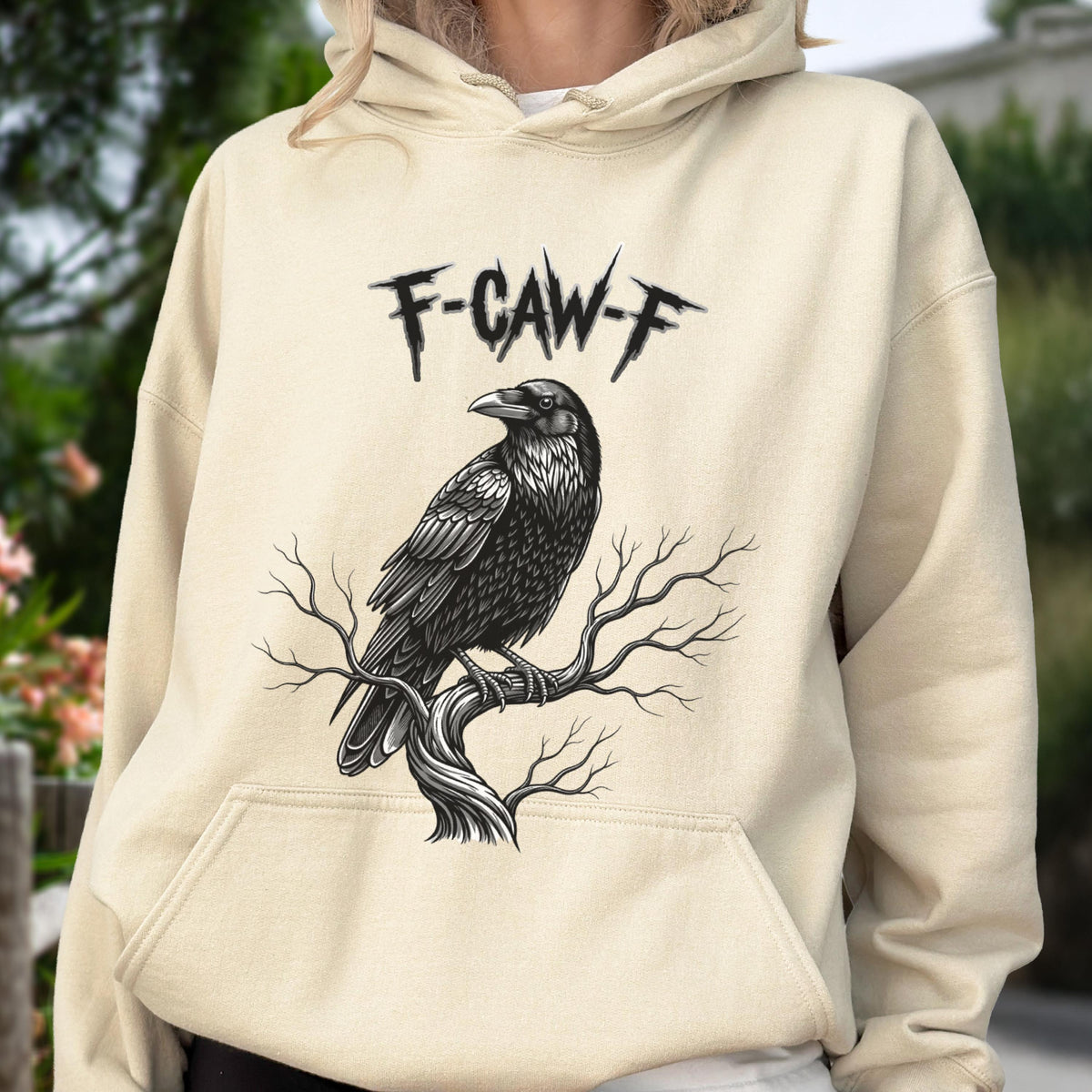 May include: A cream-colored hooded sweatshirt featuring a black graphic illustration of a raven perched on a bare tree branch. Above the bird, the text F-CAW-F is printed in a jagged, stylized black font. The garment includes a front pouch pocket.