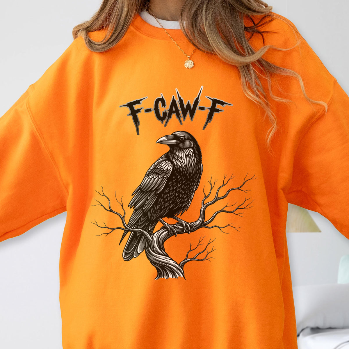 May include: A bright orange crewneck sweatshirt featuring a graphic illustration of a black raven perched on a bare, gnarled tree branch. Above the bird, the text F-CAW-F is printed in a jagged, black, stylized font across the chest area.