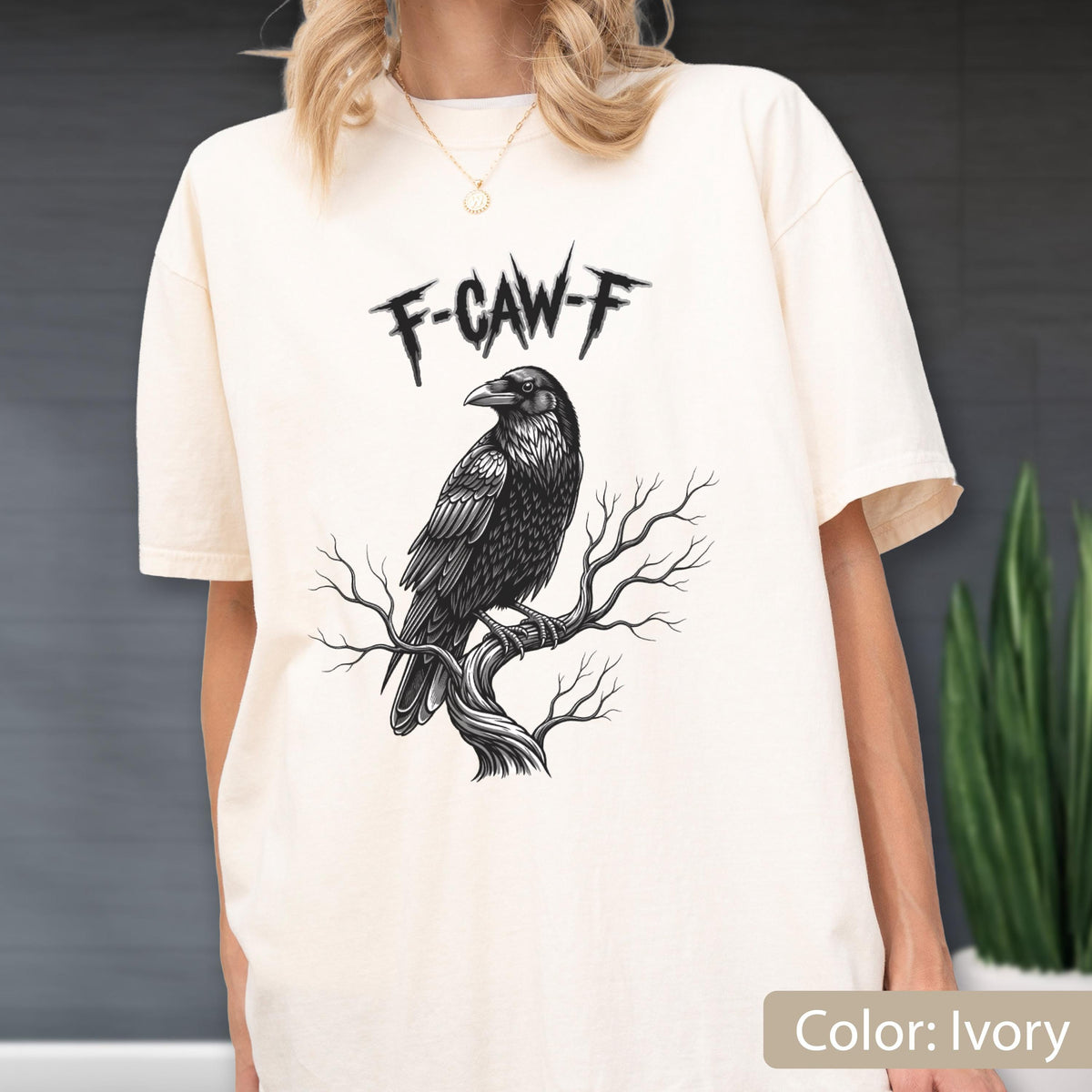 May include: An ivory short-sleeve t-shirt featuring a black graphic illustration of a raven perched on a bare tree branch. Above the bird, the text F-CAW-F is printed in a jagged, distressed black font. The shirt has a crew neckline.