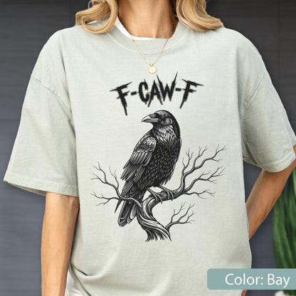May include: A bay-colored t-shirt featuring a black graphic illustration of a raven perched on a bare tree branch. Above the bird, the text F-CAW-F is printed in a jagged, stylized black font. The shirt has a crew neckline and short sleeves.