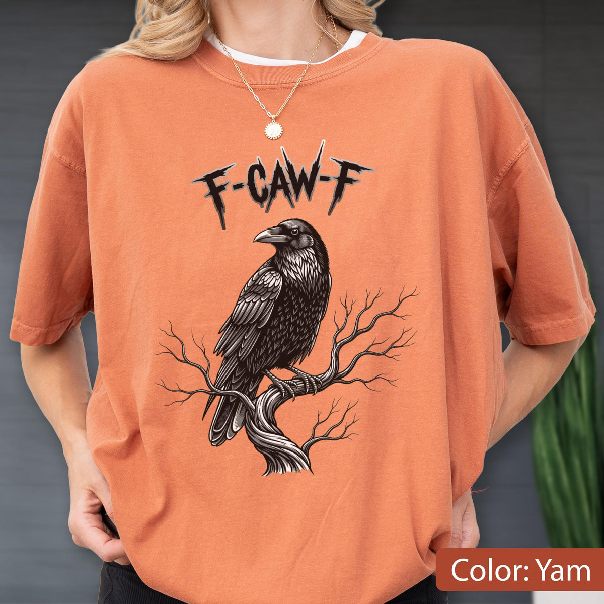 May include: A yam-colored crewneck t-shirt featuring a graphic of a black raven perched on a bare tree branch. Above the bird, the text F-CAW-F is printed in a jagged, black, stylized font. The shirt has short sleeves and a relaxed fit.
