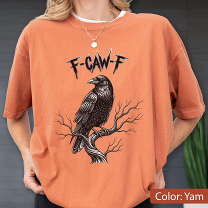 May include: A yam-colored crewneck t-shirt featuring a graphic of a black raven perched on a bare tree branch. Above the bird, the text F-CAW-F is printed in a jagged, black, stylized font. The shirt has short sleeves and a relaxed fit.