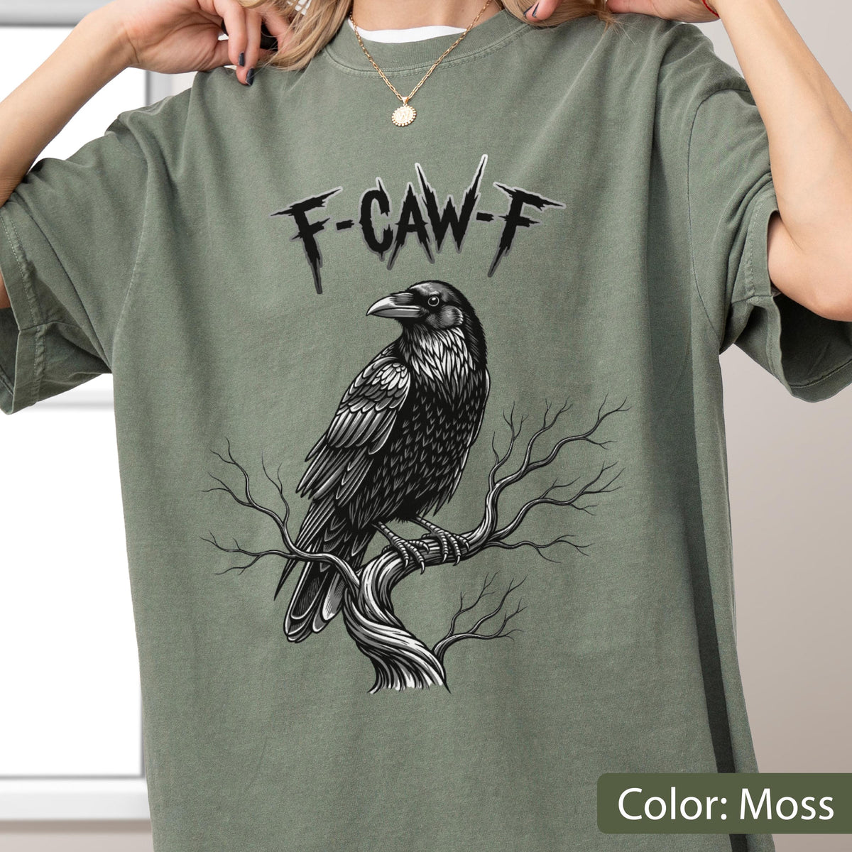 May include: A moss green short-sleeve t-shirt featuring a black and white graphic of a raven perched on a bare tree branch. Above the bird, the text F-CAW-F is printed in a jagged, stylized black font. The shirt has a crew neckline.