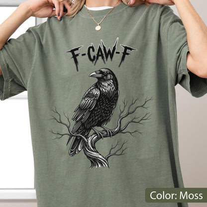 May include: A moss green short-sleeve t-shirt featuring a black and white graphic of a raven perched on a bare tree branch. Above the bird, the text F-CAW-F is printed in a jagged, stylized black font. The shirt has a crew neckline.