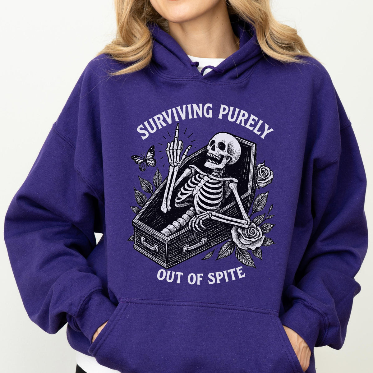 May include: A dark purple hoodie with a graphic of a skeleton in a coffin, giving the middle finger. The text reads "SURVIVING PURELY OUT OF SPITE". The design includes a butterfly and roses.