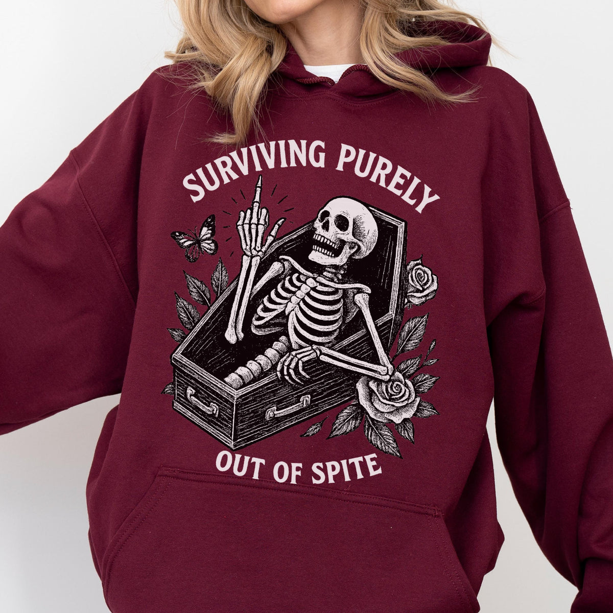 May include: Burgundy hoodie featuring a skeleton in a coffin, giving the middle finger. The graphic includes the text "SURVIVING PURELY OUT OF SPITE" with a butterfly and roses. The hoodie is a comfortable, casual style.