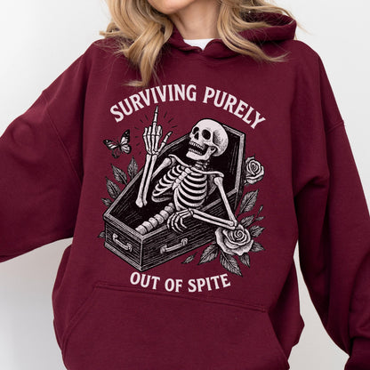 May include: Burgundy hoodie featuring a skeleton in a coffin, giving the middle finger. The graphic includes the text "SURVIVING PURELY OUT OF SPITE" with a butterfly and roses. The hoodie is a comfortable, casual style.